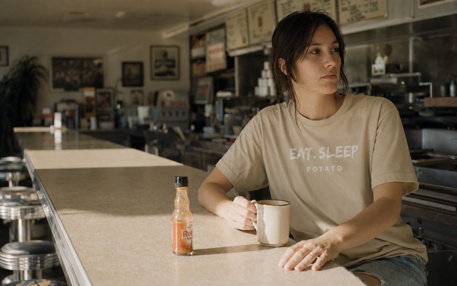 A pledged spud in an Eat Sleep Potato tee at a diner counter, coffee and hot sauce within reach.